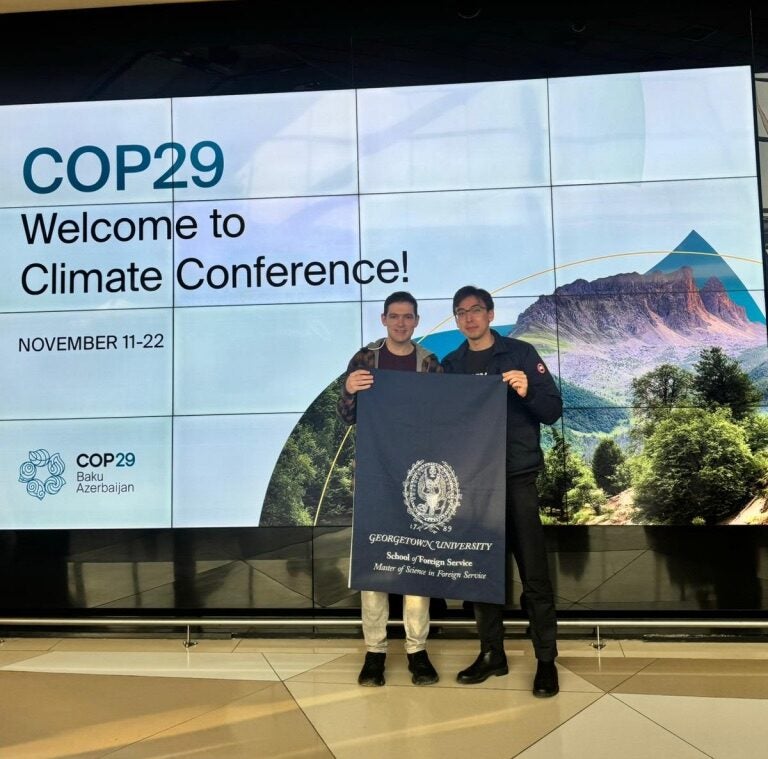 Zack Slotkin (MSFS'25) and Kamal Aubakirov (MSFS'25) pictured holding a Georgetown University banner in front of a COP29 welcome sign.